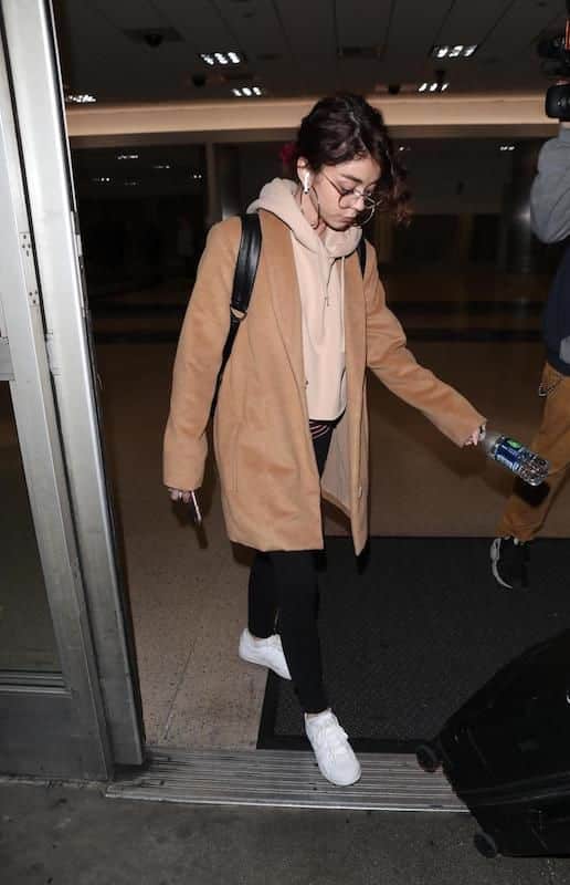 Sarah Hyland wearing a nude cropped hoodie, a camel colored mid-length coat, black leggings, hoop earrings, a black leather backpack, and white low-top sneakers