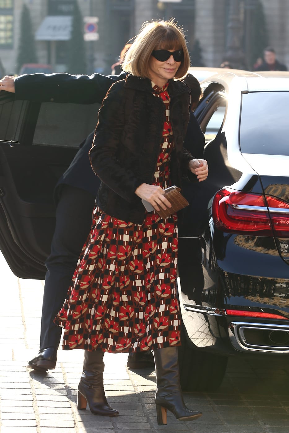 Anna Wintour wearing a red patterned dress, black boots, and a puffy coat