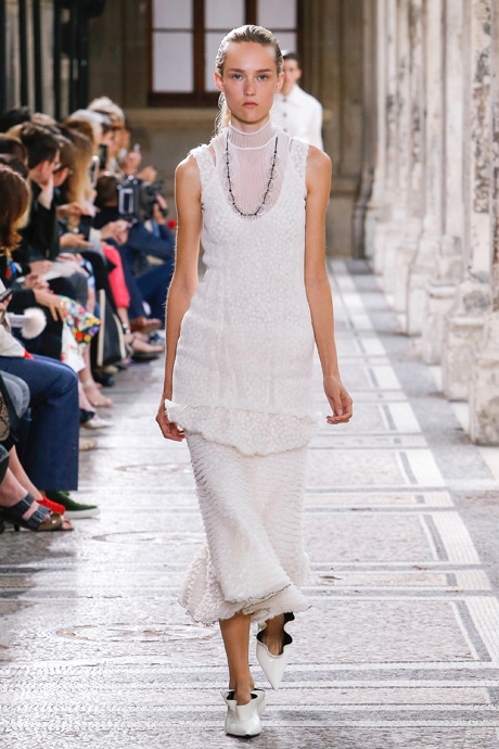 Proenza Schouler Spring 2018 model on the runway wearing an all white outfit, including a long white skirt with ruffles, a sheer white sleeveless layered tunic top, a beaded statement necklace, and white patent ruffle flats with pointed toes