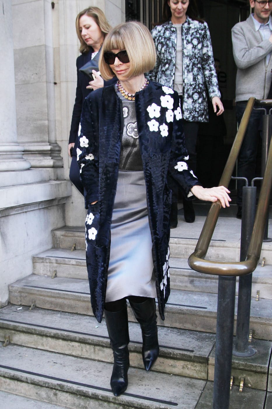 Anna Wintour wearing a gray satin pencil skirt, a gray floral top, a black and white floral fur coat, and a statement necklace with boots