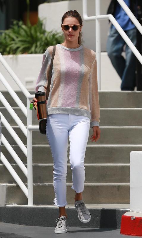 Alessandra Ambrosio wearing a multicolor striped crewneck sweater, white cropped ankle jeans, beige frame sunglasses, and gray low-top sneakers