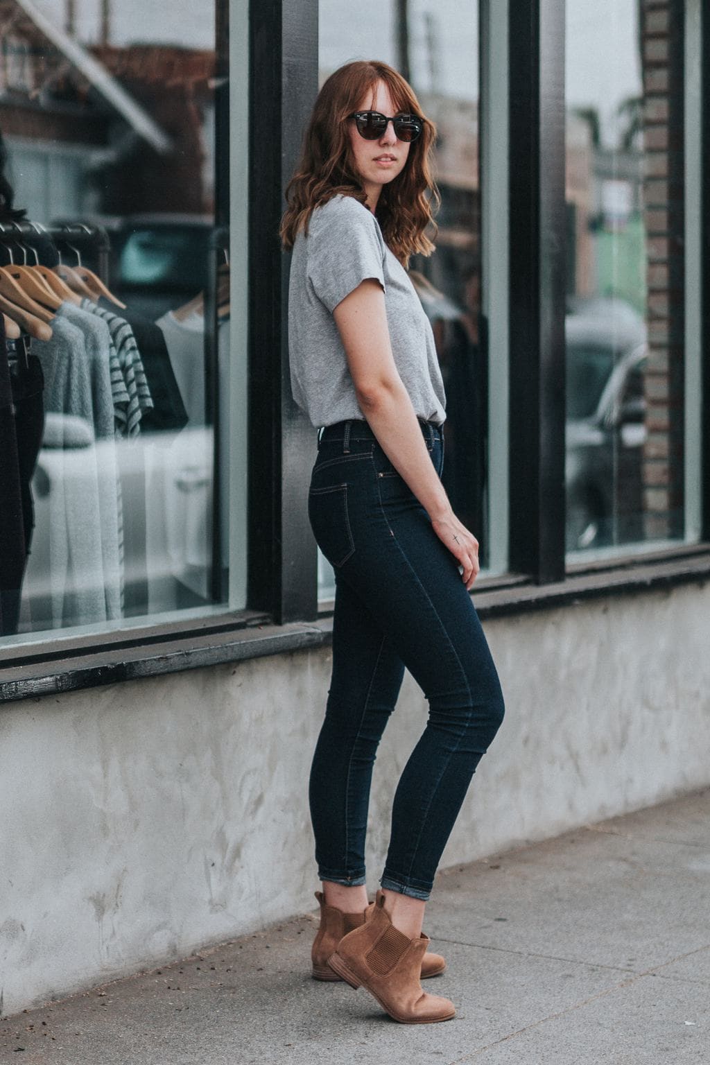 Photo of woman standing to the side with her face to the camera wearing sunglasses, gray short-sleeved t-shirt tucked into dark-wash skinny jeans, and light brown Chelsea boots