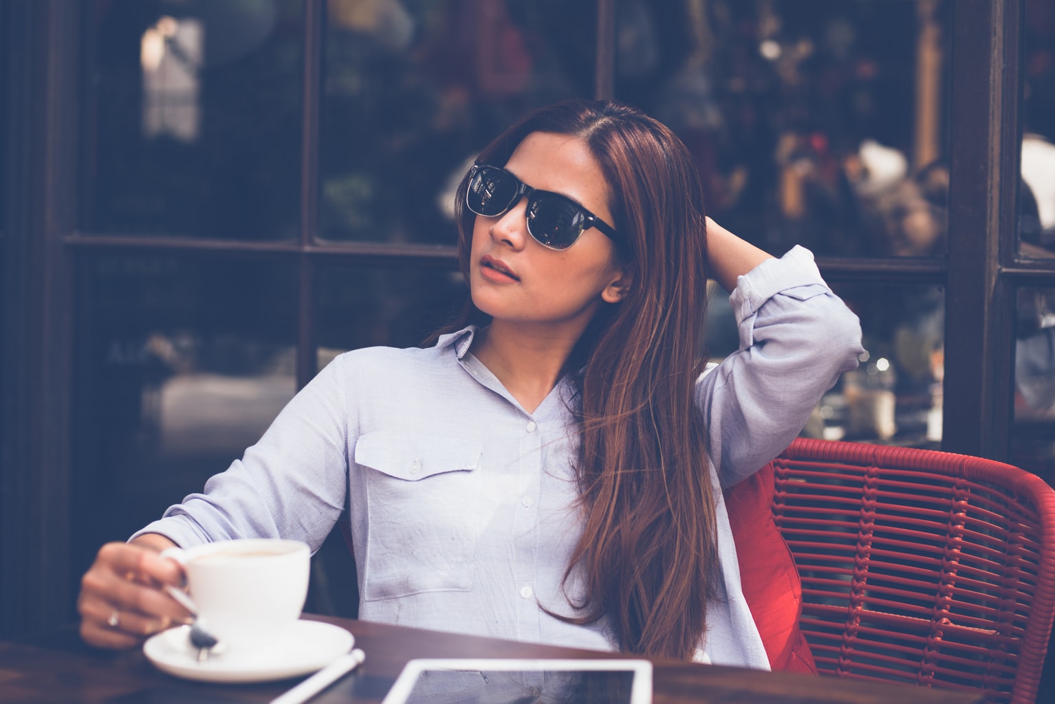 Photo of a young woman wearing sunglasses and a button-up shirt, sitting at a table in a cafe holding a coffee cup