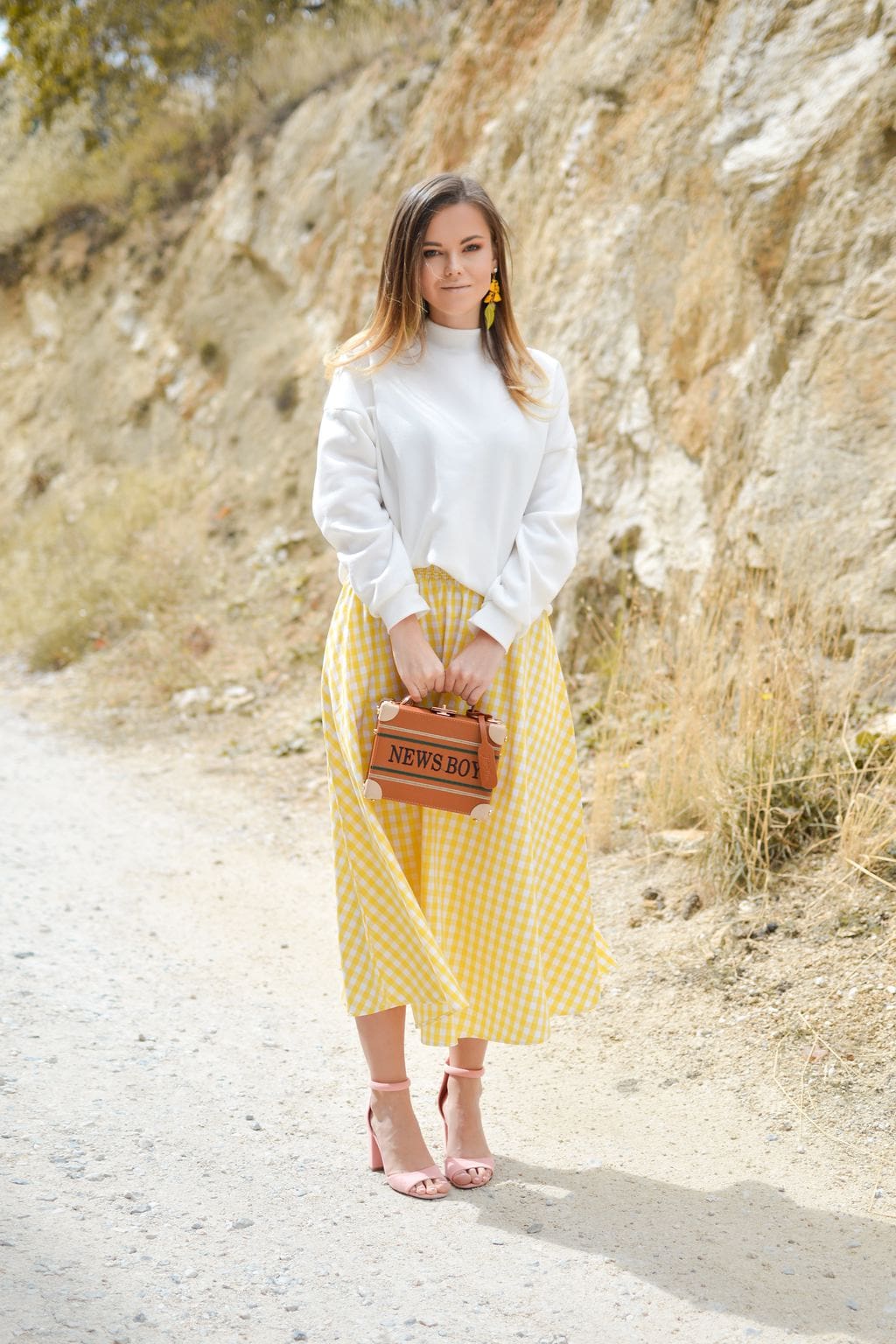 Photo of woman standing against rocky background wearing yellow dangling earrings, white turtleneck top, yellow and white gingham skirt, and pale pink heeled sandals, holding a cognac brown box bag that says 