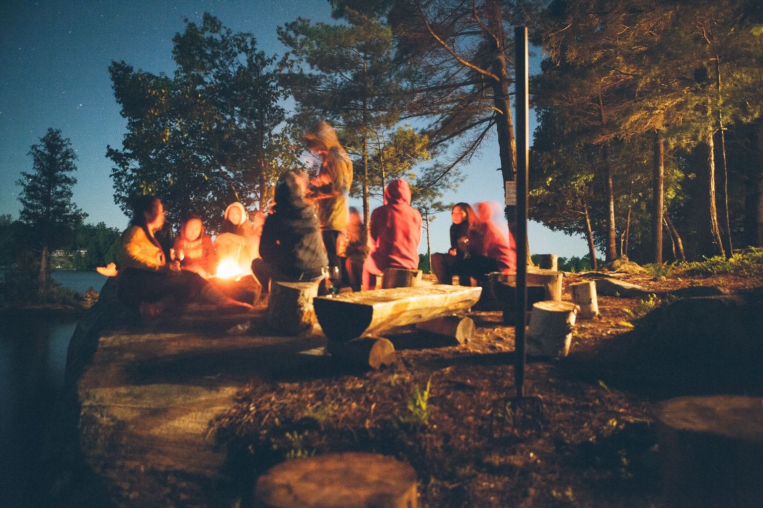 Photo of a group of people sitting around a campfire in the woods by the water at twilight