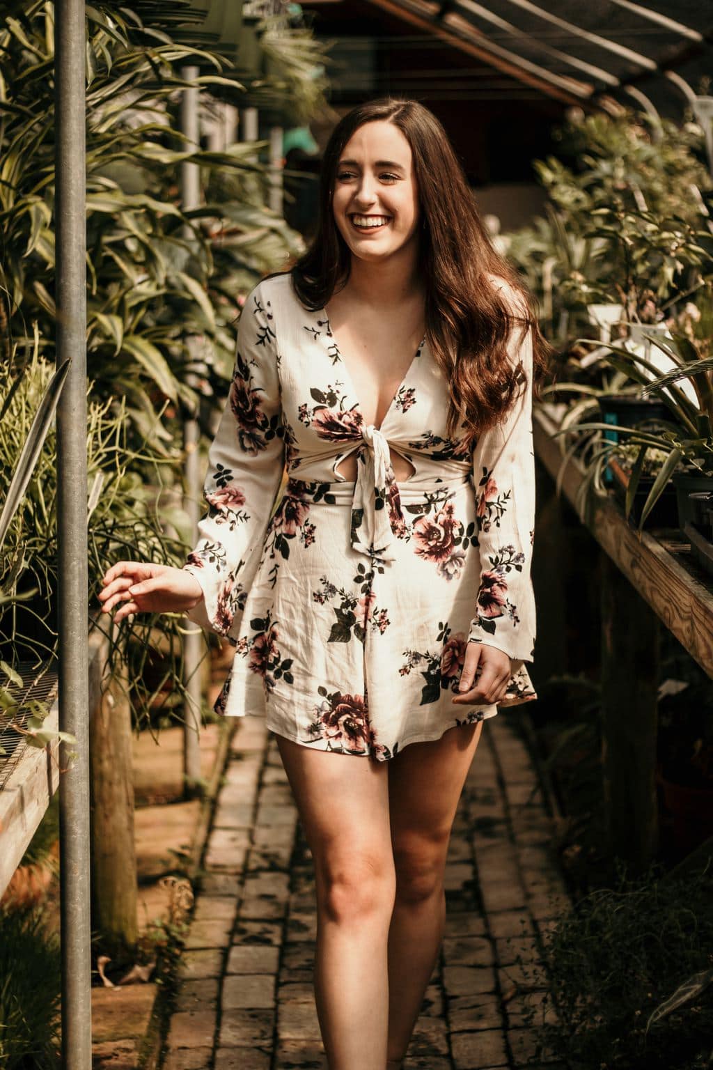 Woman, walking in a greenhouse, wearing a long-sleeved off-white romper with a pink and green floral print and open tie front