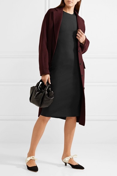 Woman wearing black sheath dress, maroon coat, and black pointed-toe kitten-heel mules with white bow, and carrying small black leather bag