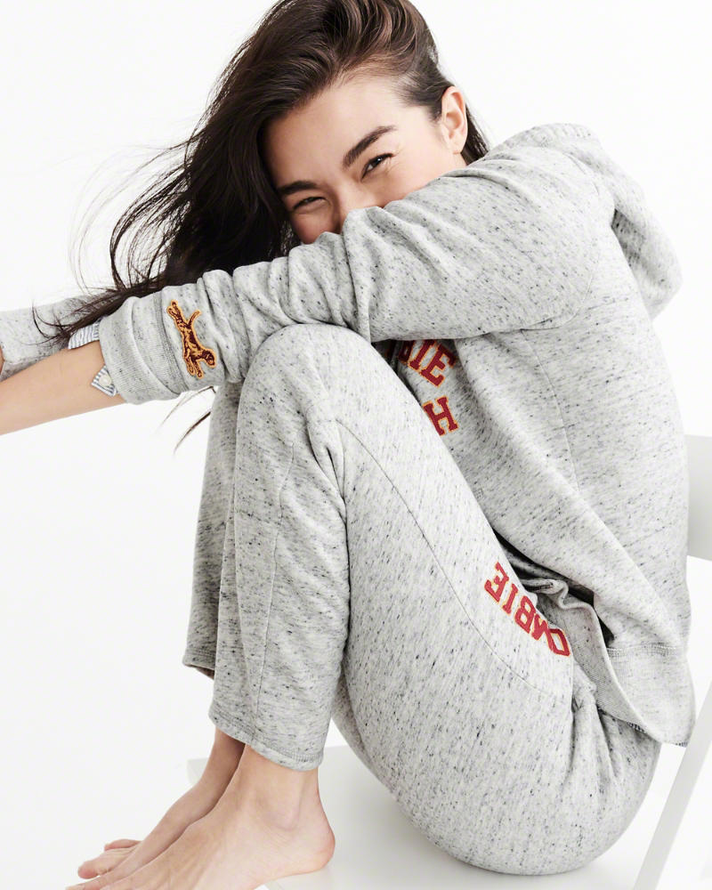 Woman sitting down on a white chair with her knees drawn close to her body wearing matching light heather gray sweatpants and sweatshirt with Abercrombie & Fitch logo in red outlined in yellow and dog on sweatshirt sleeve, with the sleeve of a blue-and-white button-up shirt peeking out from under one of the arms of the sweatshirt