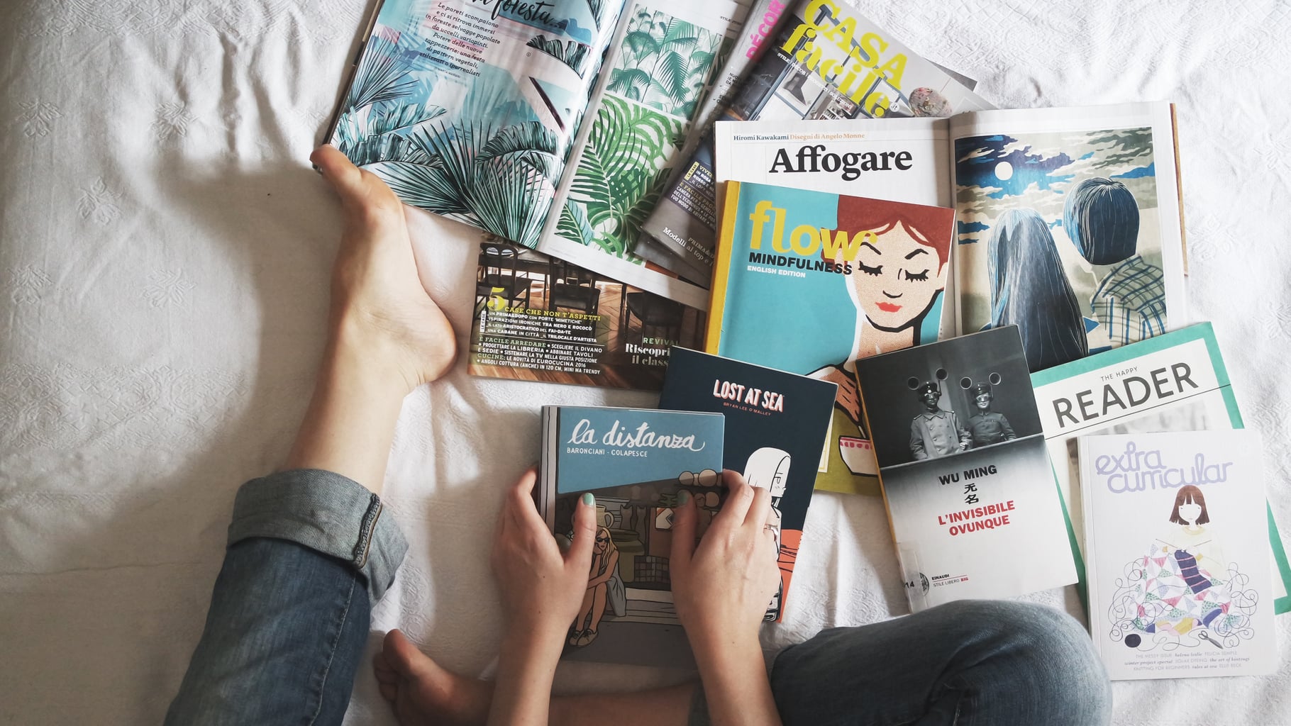 Legs wearing jeans on a bed with white sheets and a pile of colorful books of different types.