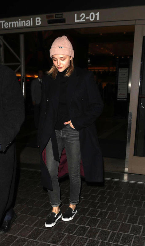 Chloe Moretz wearing a blush pink knit beanie hat, black sweater, long black coat, gray straight leg jeans, and black Vans slip on sneakers