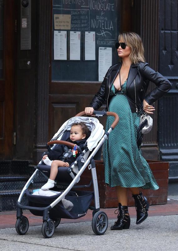 Chrissy Teigen wearing a black leather moto jacket, a green and white v-neck polka dot midi dress, black shield sunglasses, a silver and black tassel crossbody bag, and black leather lace-up booties