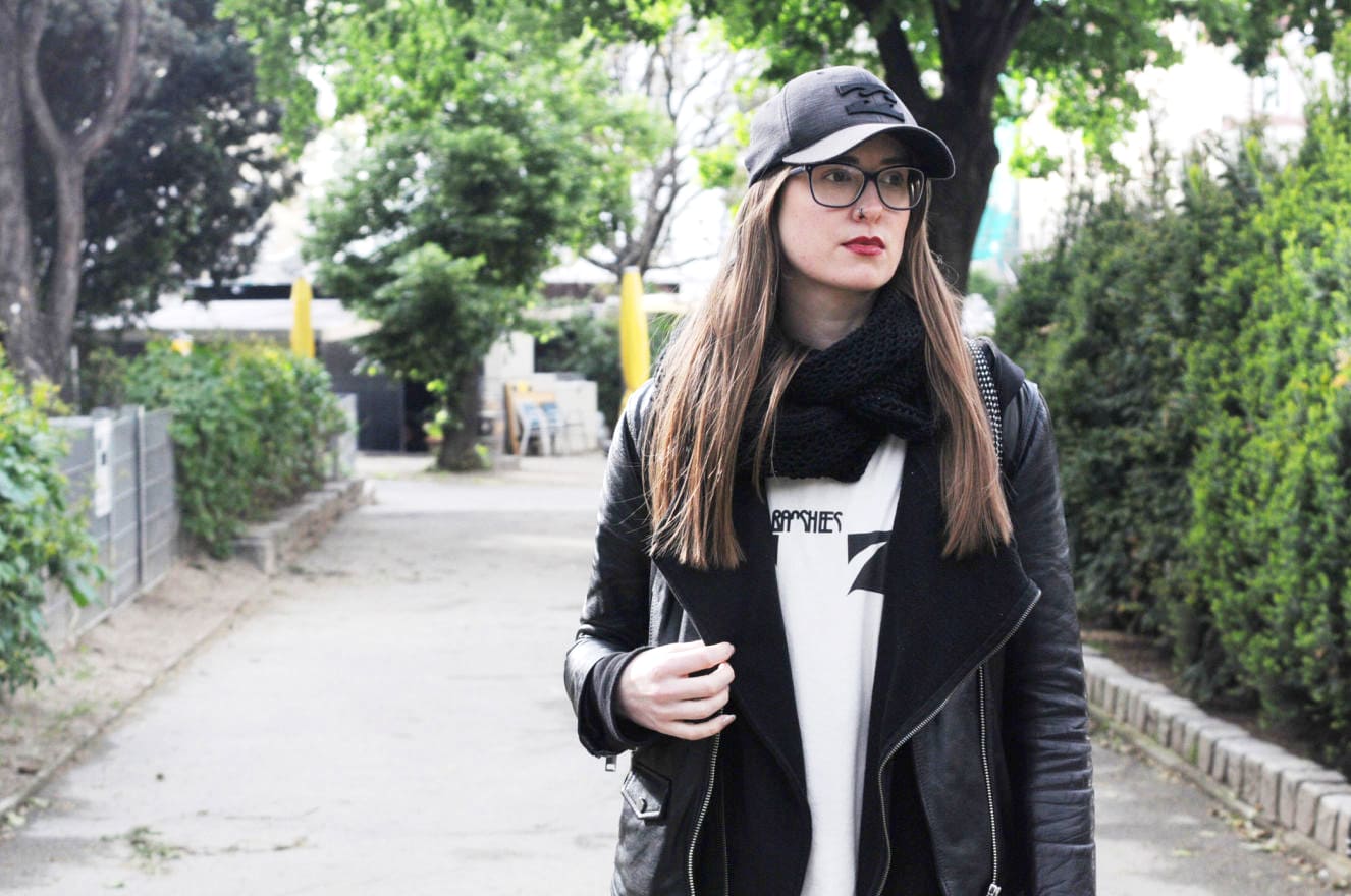 College student at University of Vienna wearing an edgy style black leather jacket, band tee shirt, hoodie, glasses, red lipstick and baseball cap