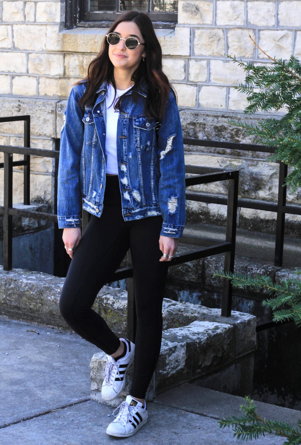 Kansas State University student Bailey wears a casual white tee with high-rise black leggings, a distressed, oversized denim jacket, worn-in Adidas all star sneakers, and round retro gold-rimmed sunglasses.