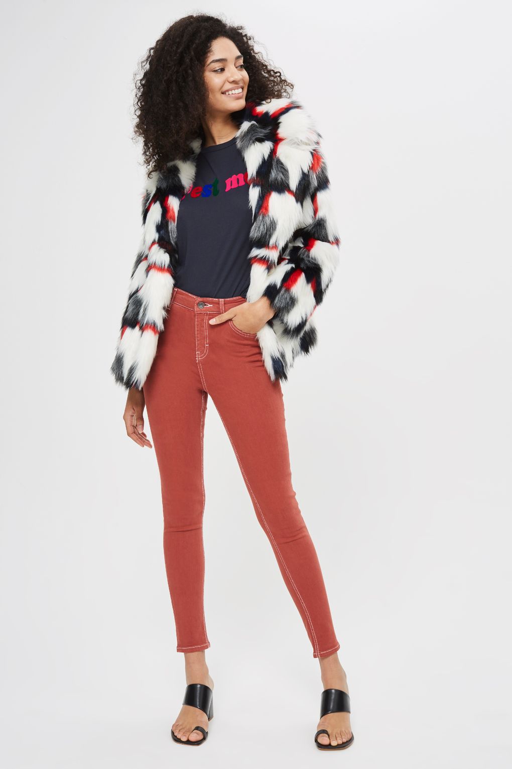 Woman wearing white, black, red, and gray faux-fur jacket, black t-shirt with multi-colored "C'est moi" graphic, terracotta-colored skinny jeans with white stitching, and black sandals with wide strap and toe-ring