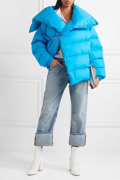 Woman wearing bright blue oversized puffer coat with safety-pin closure over rolled-up jeans and striped button-up, with white patent block-heeled ankle boots and white oversized clutch