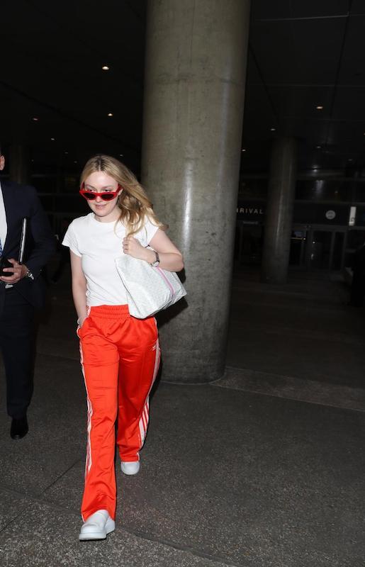 Dakota Fanning wearing red cat-eye sunglasses, a white t-shirt, red and white striped track pants, white sneakers, and a white shoulder bag
