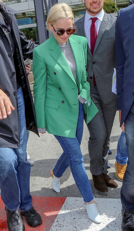 Emilia Clarke wearing a green double-breasted oversized blazer, a gray mock neck top, round tortoise sunglasses, medium wash straight leg jeans, and wedge mules