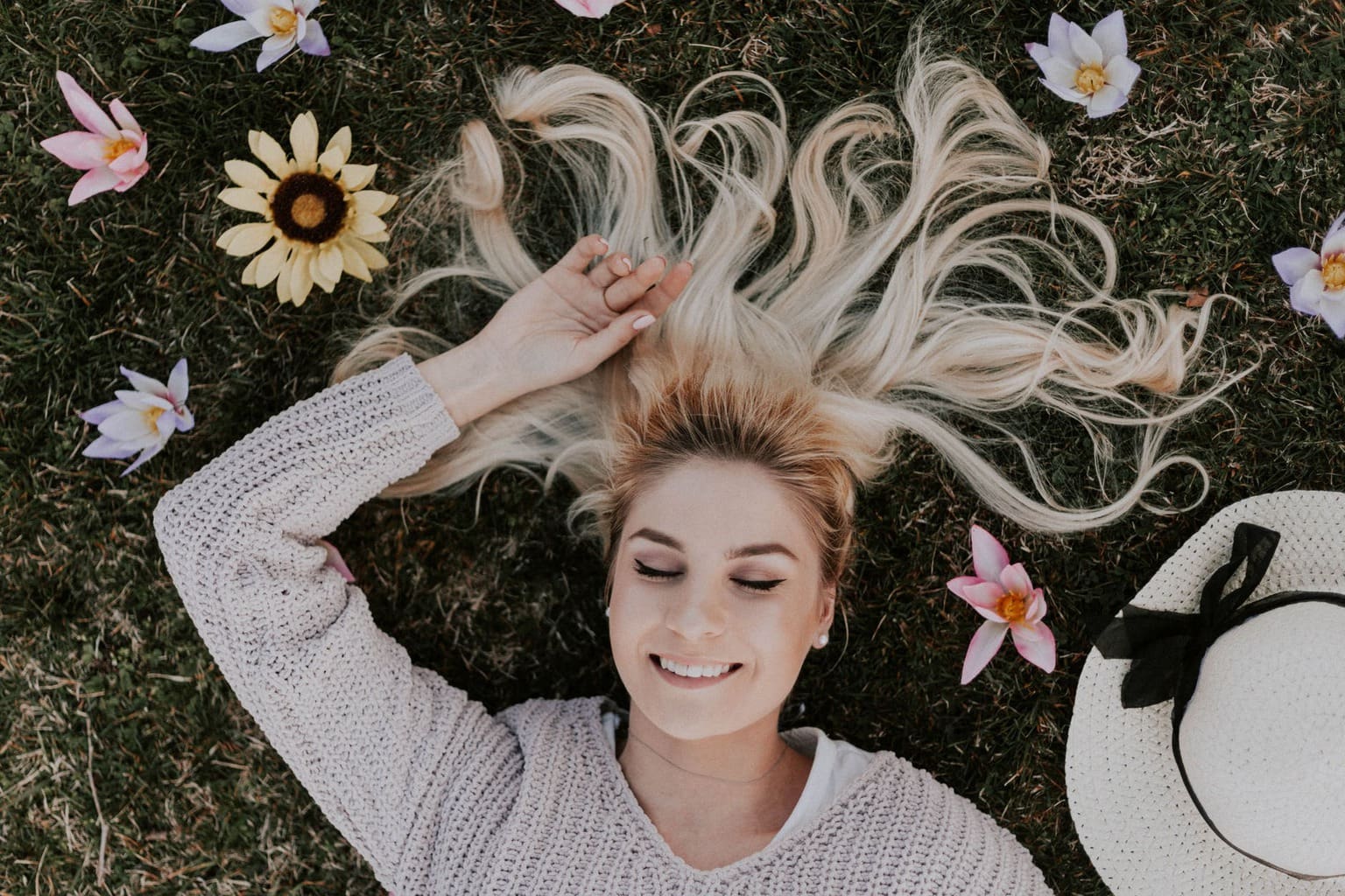 Blonde girl sitting in grass with hair laid out, flowers, and a hat