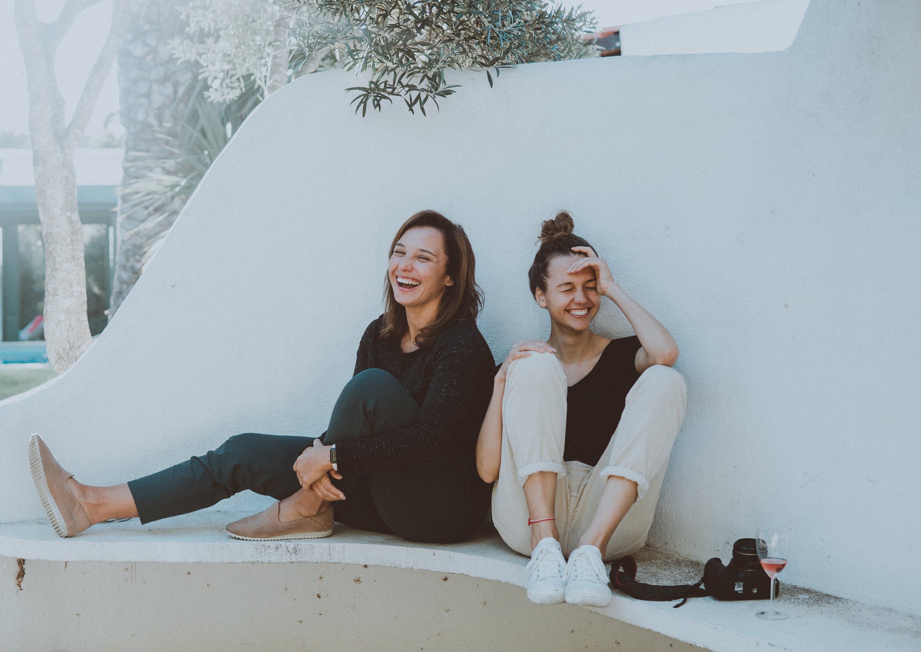 Two woman sitting on a bench laughing with wine and a camera