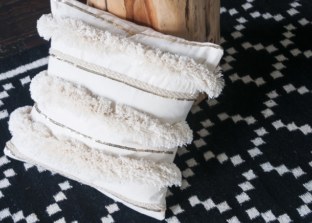 Photo including a fringe pillow leaning against a wooden table sitting on a geometric rug.