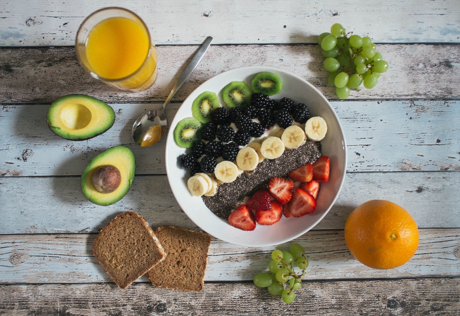 variety of fruits (kiwi, grapes, avocado, orange banana, strawberry)  in a bowl with orange juice and bread on a wooden table