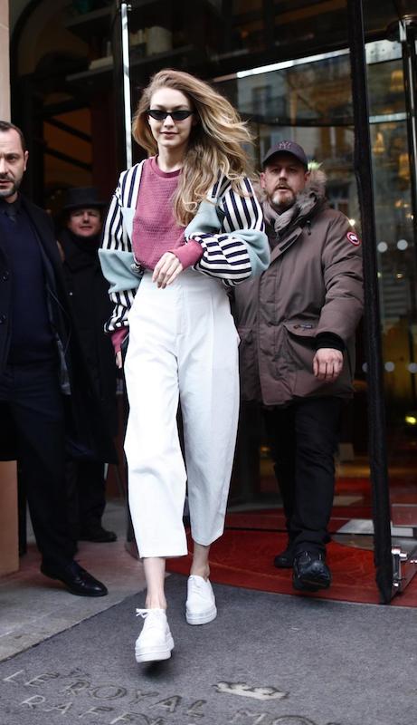 Gigi Hadid wearing black cat-eye sunglasses, a multicolor striped top, white high-waist cropped pants, and white platform lace-up shoes