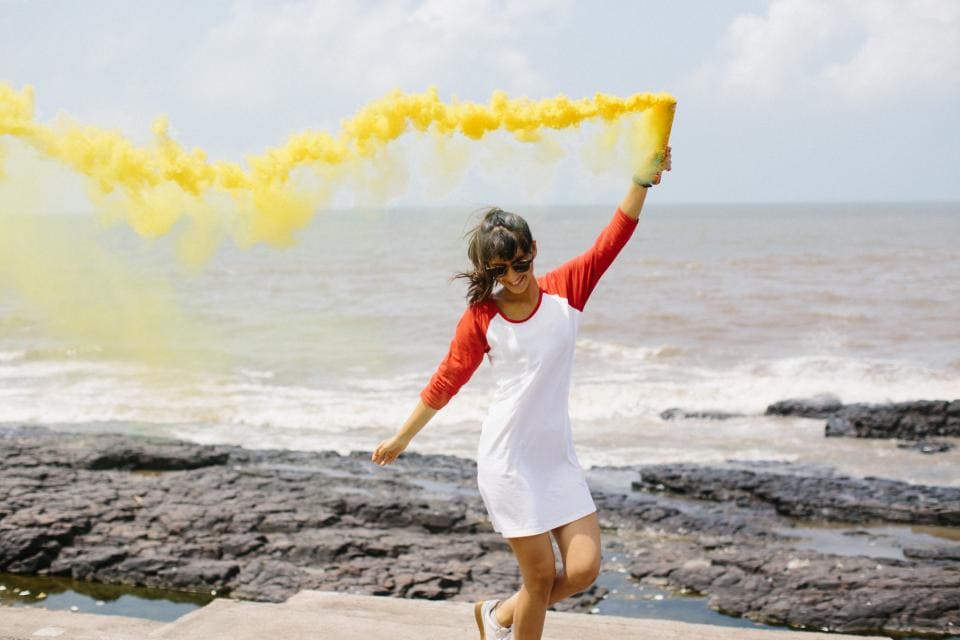 Photo of girl running with yellow chalk dust in her left hand. She's next to the beach and wearing a red and white henley dress and white sneakers.