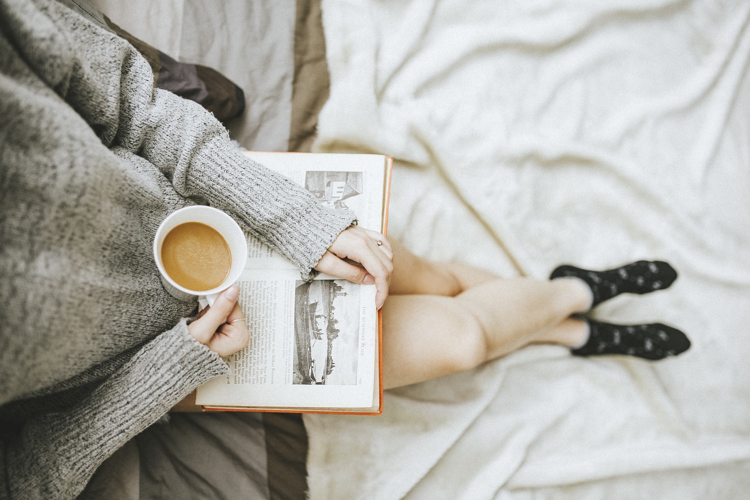 Photo of girl wearing a gray sweater and black patterned socks reading a book and drinking coffee.