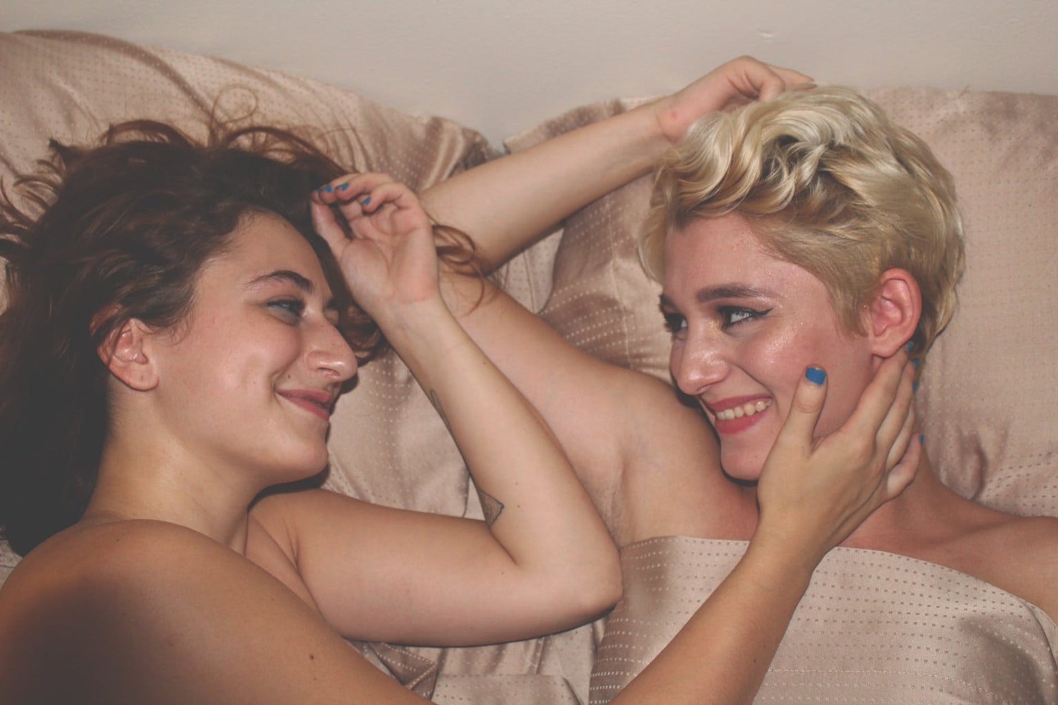 Girl with brown hair in bed with girl with short blonde hair with half on face