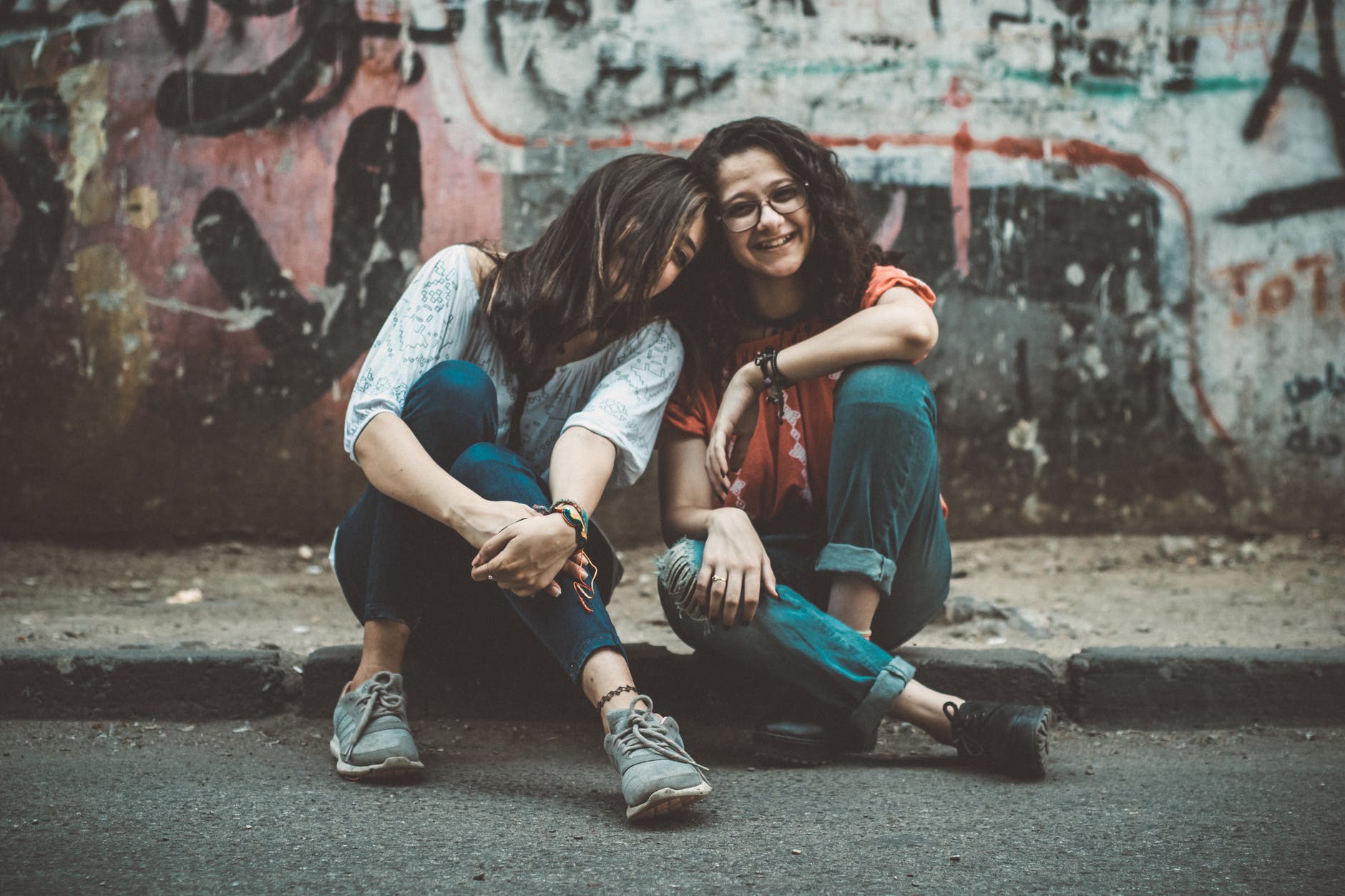 Two girls sitting on the street with graffiti laughing