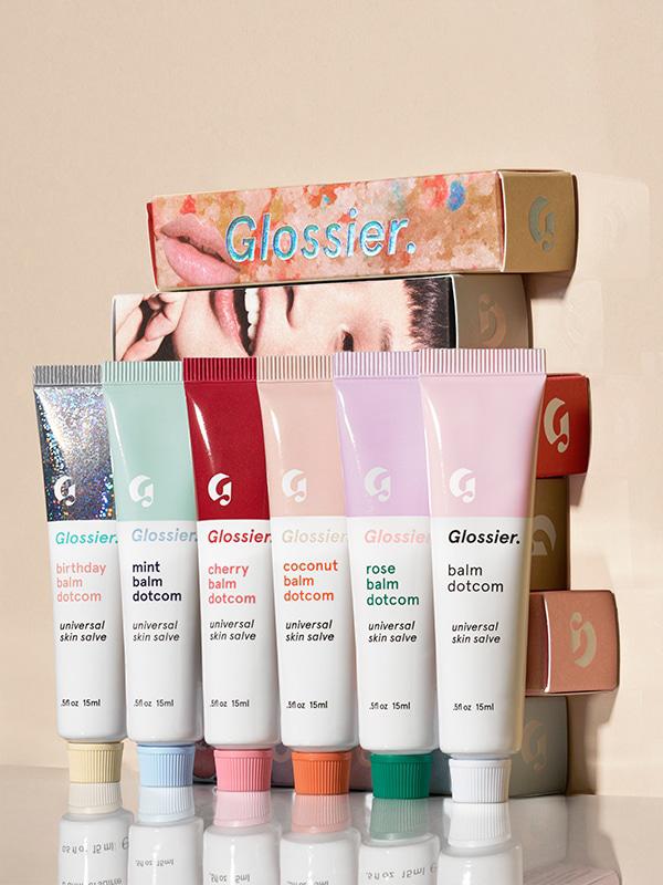 Photo of all Glossier Balm Dotcom flavors (Birthday Balm Dotcom, Mint Balm Dotcom, Cherry Balm Dotcom, Coconut Balm Dotcom, Rose balm Dotcom, Original Balm Dotcom) in front of boxes of the product stacked up on a beige surface against a beige background