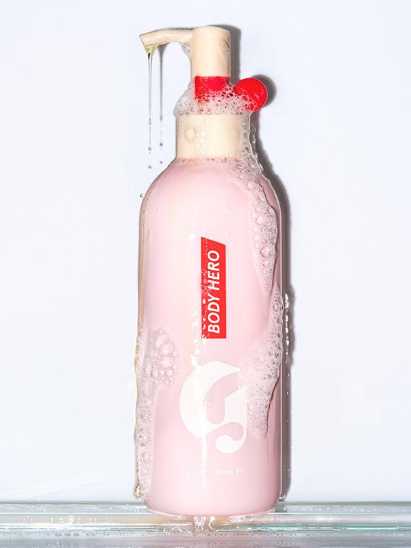 Photo of Glossier Body Hero Daily Oil Wash on glass shelf against light gray background, with body wash coming out of the pump and suds covering the bottle