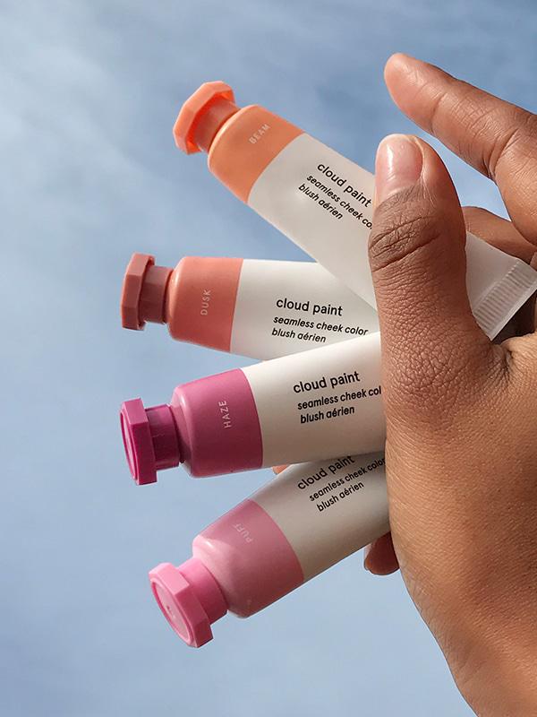Close-up photo of hand holding four tubes of Glossier Cloud paint blush in a vertical fan-like formation in the shades Beam, Dusk, Haze, and Puff, against a backdrop of blue sky with wispy clouds