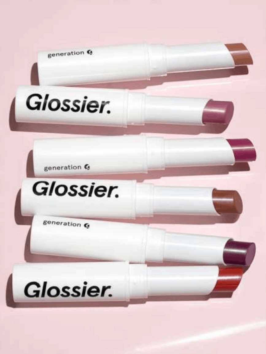 Tubes of Glossier Generation G lip color laid down against a glossy pale pink background, in the shades (from top to bottom): Cake, "a subtle peachy nude"; Like, "a light powdery pink"; Crush, "a hot raspberry pink"; Leo, "a warm cocoa brown"; Jam, "a deep berry magenta"; and Zip, "a poppy red."