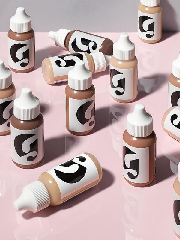 Close-up photo of upright and sideways containers of Glossier Perfecting Skin tint in all shades (Light, Medium, Dark, Deep, Rich) on a glossy, reflective pale pink surface, against a grey-ish background