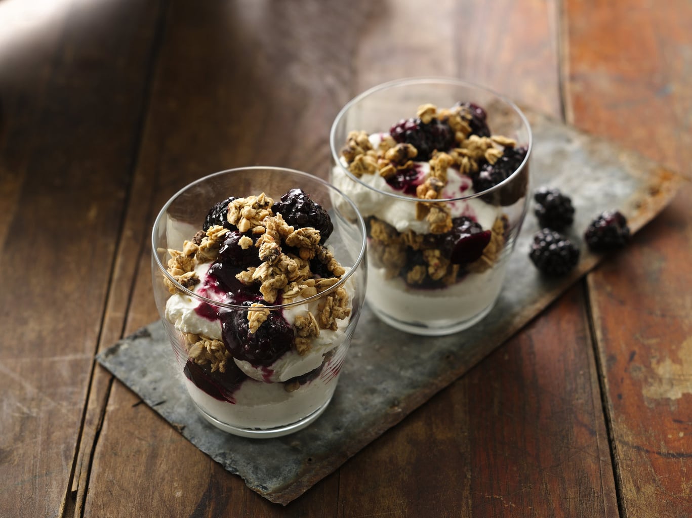 Two blackberry and granola parfaits sit on a tray on a wooden table.