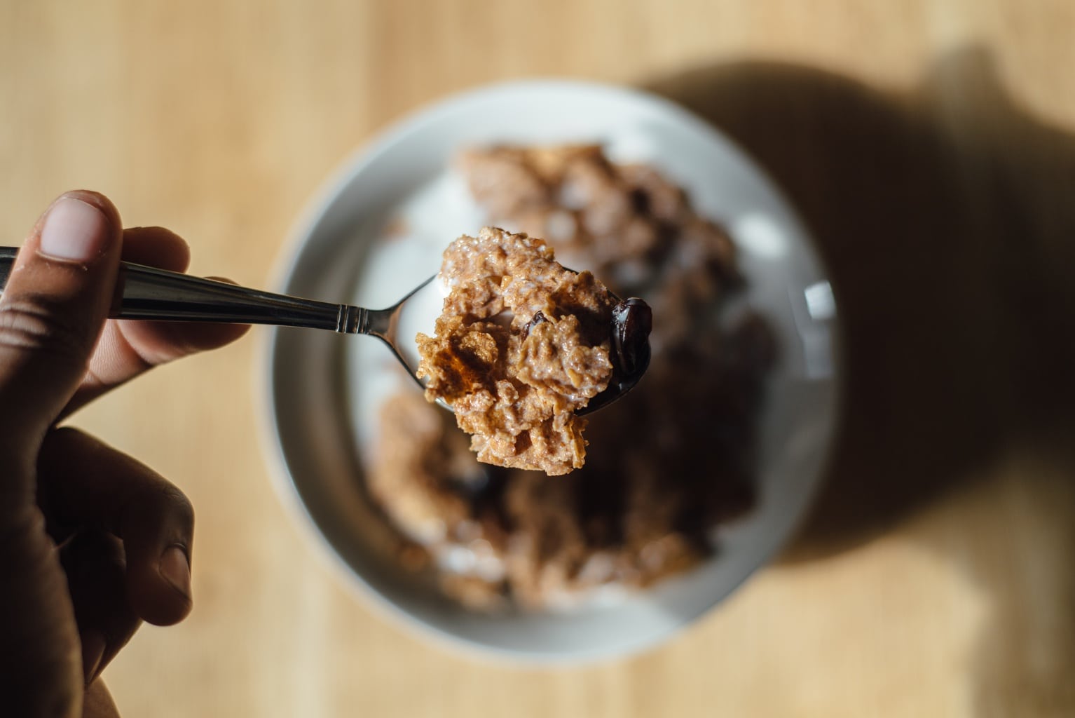 A hand holds a spoonful of cereal over a full bowl sitting on a table.