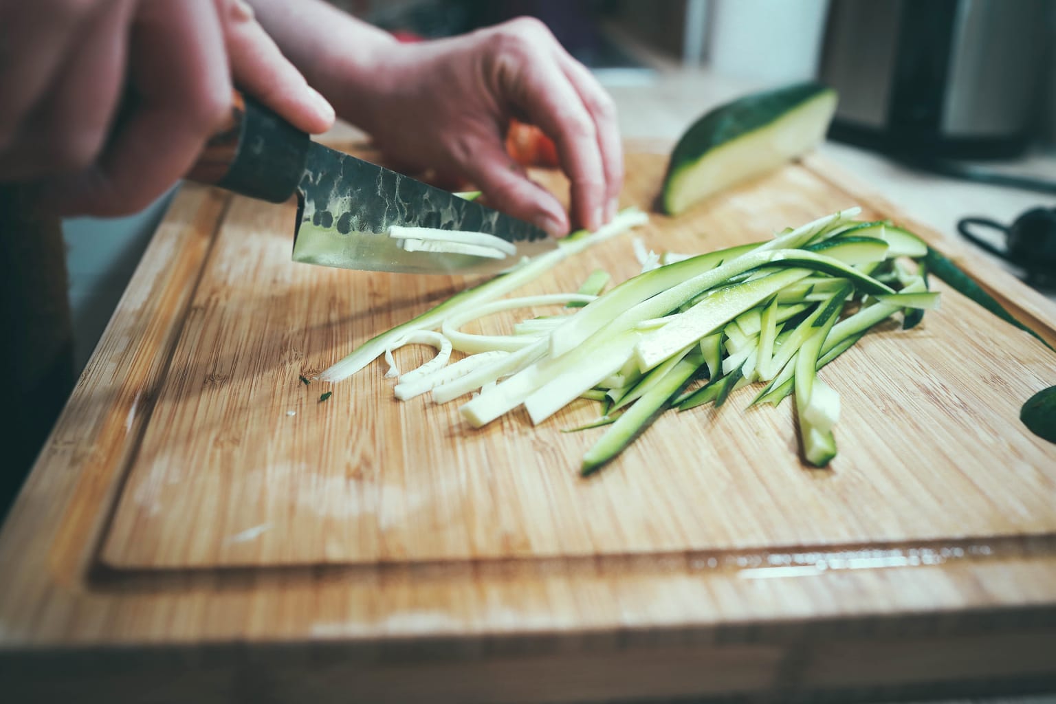 A pair of hands use a kitchen knife to slice thin strips of cucumber on a cutting board.