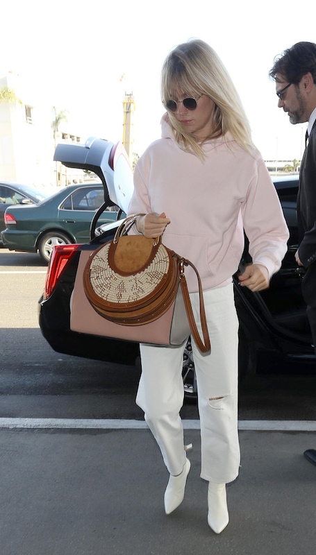 January Jones wearing a light pink hoodie sweatshirt, round sunglasses, white cropped straight leg jeans, white ankle booties, and carrying pink and brown handbags