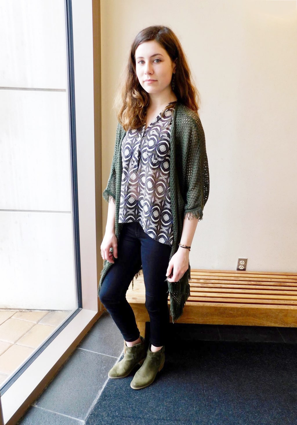 Jefferson University student Jenna sports a silky see-through black button-up blouse with a white pattern, black fitted skinny jeans, and a casual loose-knit olive green cardigan.