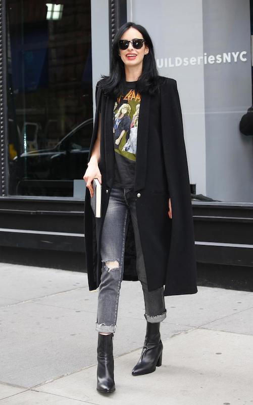 Krysten Ritter wearing a rocker print Def Leppard t-shirt, a black longline cape, dark sunglasses, gray jeans, and black leather mid-calf booties