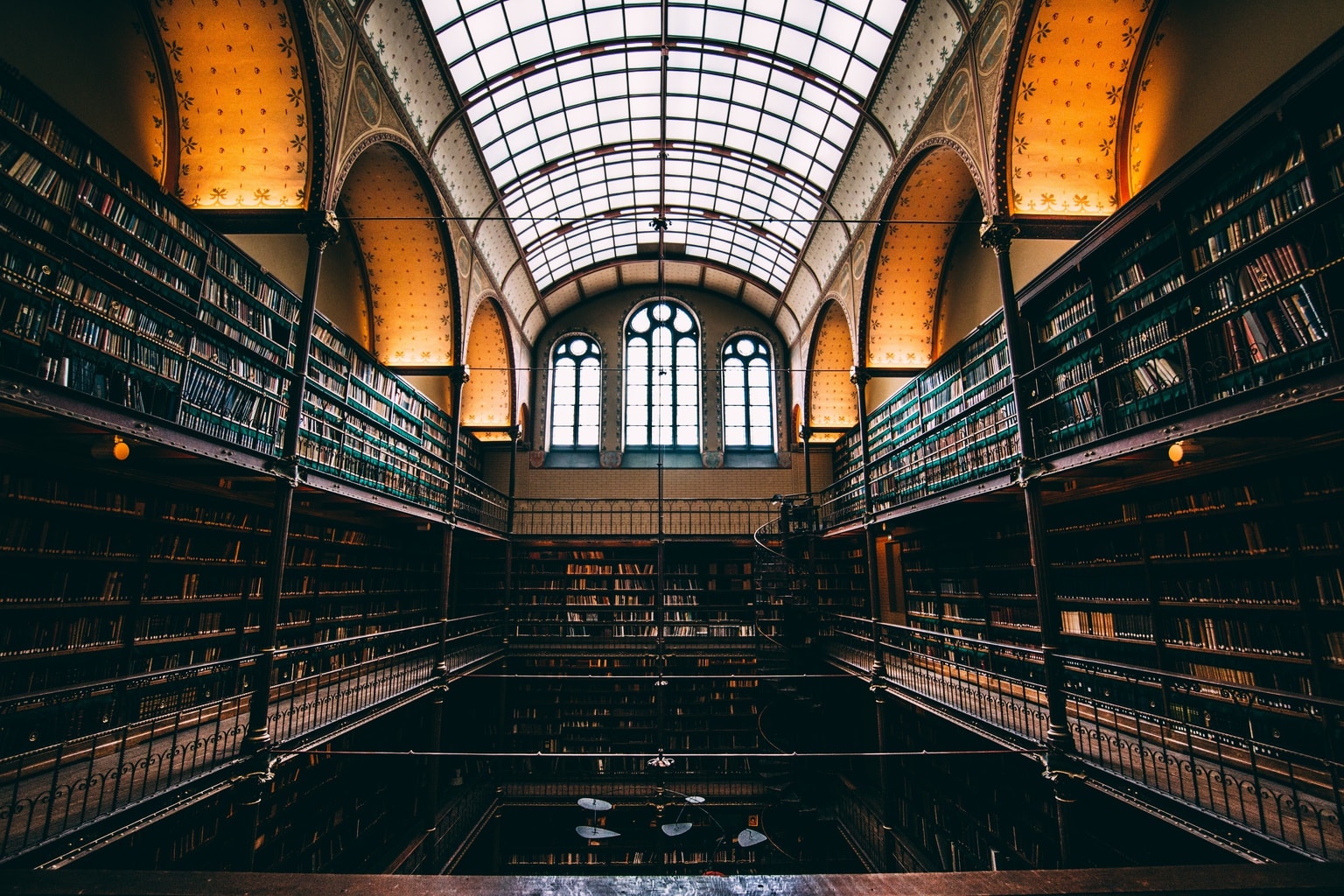 A wide view of a beautiful library with high archways, a glass ceiling and large windows surrounded by numerous shelves of books