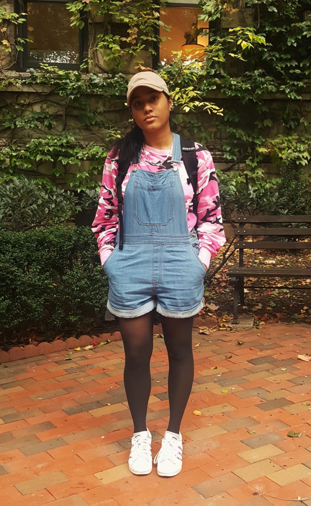 Barnard College student Alema wears a casual set of overall shorts with a bright pink camouflage long-sleeve tee underneath. She tops off her look with a tan baseball cap and black tights.