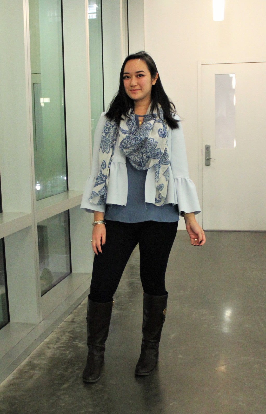 A Barnard College at Columbia University student wears a blue top with a sky blue jacket and a blue and white paisley scarf. She pairs it with black leggings and brown riding boots.