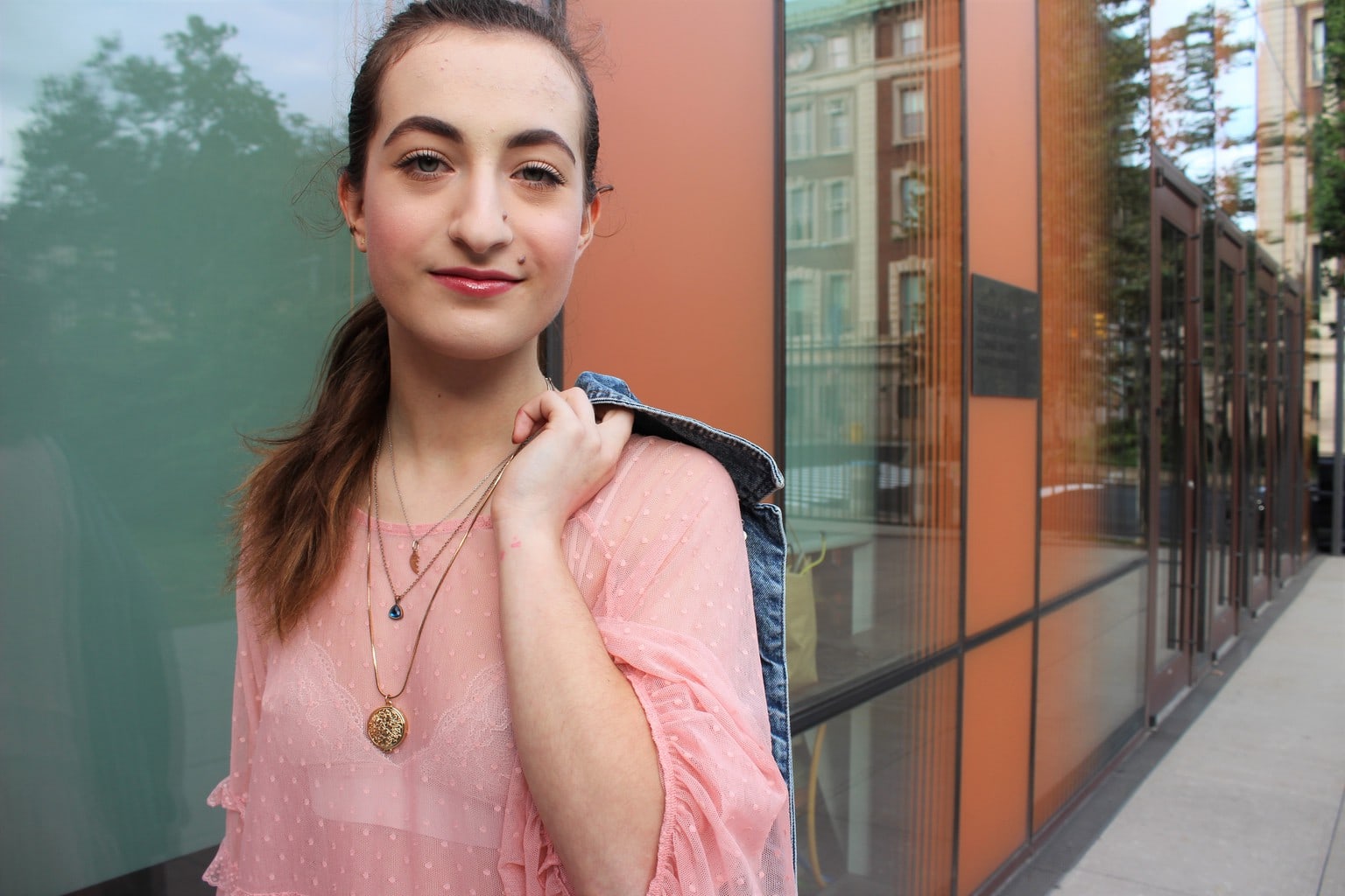 Francesca wears a sheer pink blouse with a white lace bralette underneath and accessorizes with gold layered necklaces and a light-wash denim jacket.