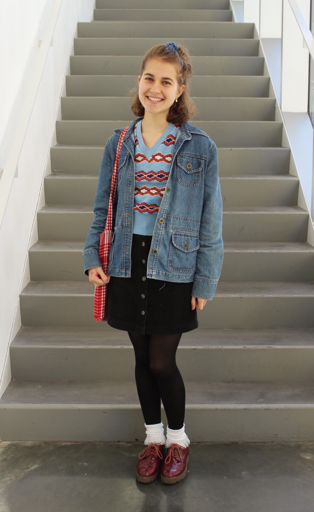 90s outfit: Barnard College at Columbia University student wears an oversized denim jacket with a baby blue and red printed v-neck sweater, a button-up black miniskirt, black opaque tights, and ruffled white socks with chunky maroon lace-up shoes.