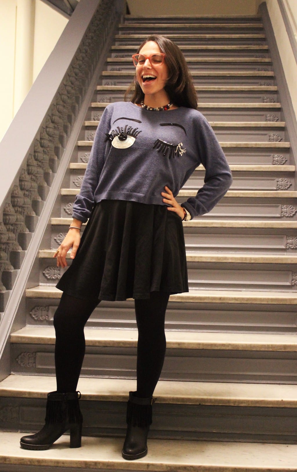 This Barnard College student wears a grey winking eye handmade grey sweater with a flowy black skirt with opaque black tights and fringe heeled black booties. She accessorizes with layered beaded necklaces and bright red eyeglasses.