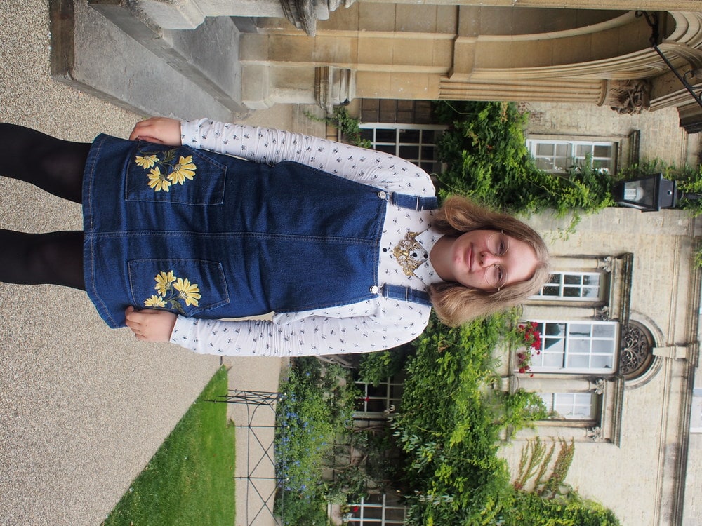 Hannah, a student at the University of Oxford, wears a denim jumper dress with yellow embroidery on the pockets, white patterned button-up with a collar, and a gold statement necklace.