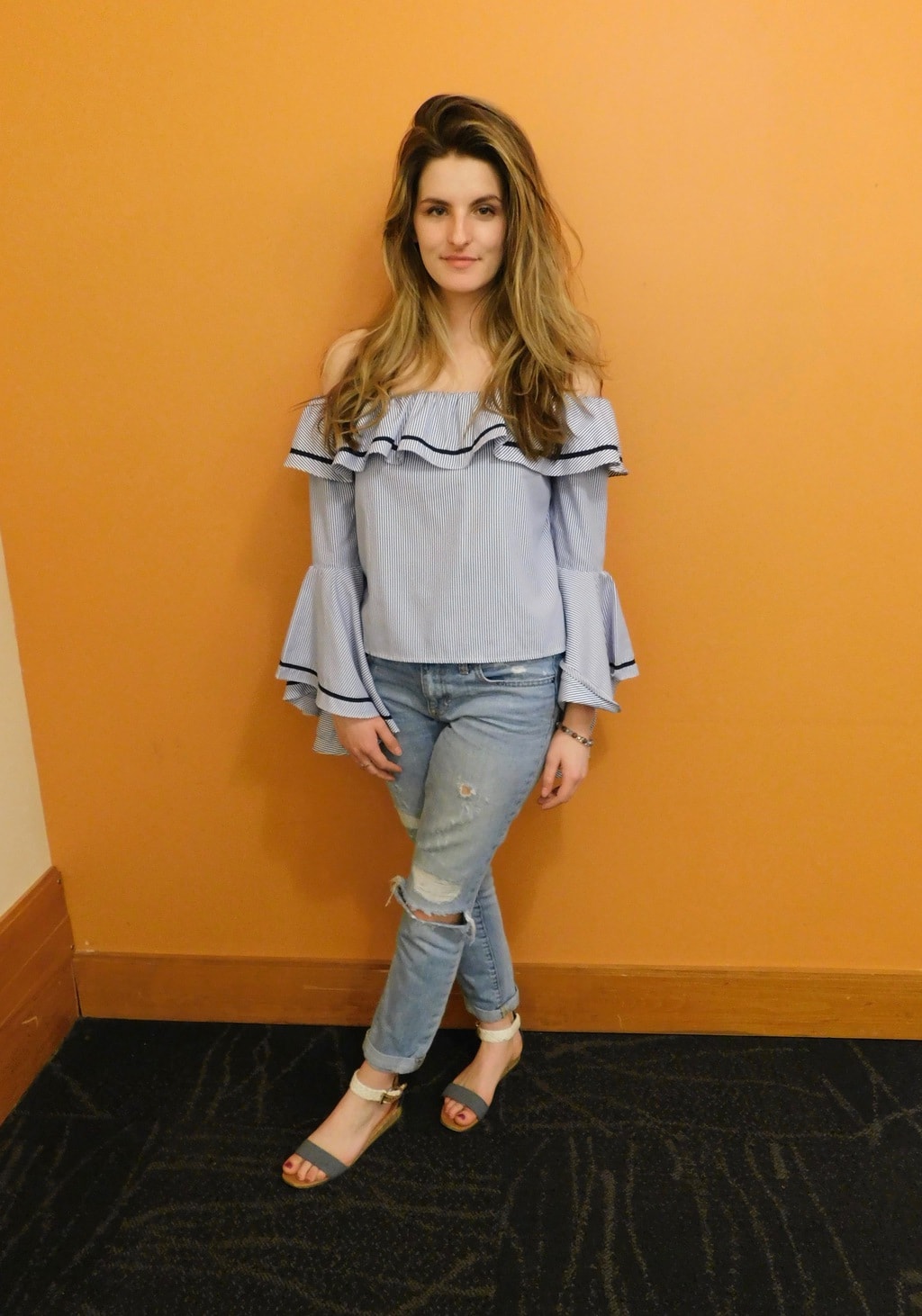 Jefferson University student Danielle wears an off-the-shoulder blue ruffled top with bell sleeves, light-wash distressed denim jeans, and simple grey and white flat sandals.