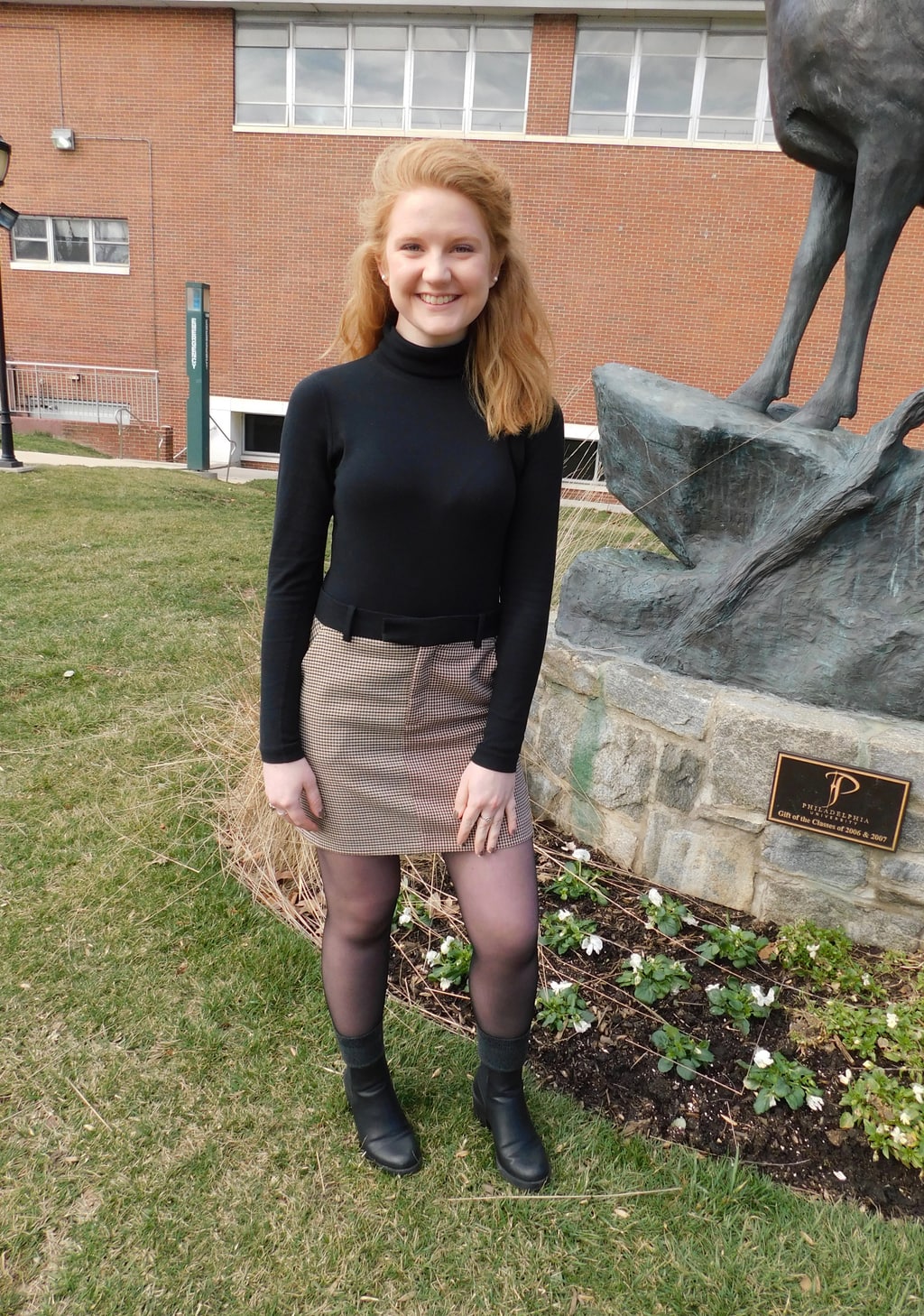 Hannah, a student a Jefferson University, wears a black long-sleeve turtleneck sweater tucked into a maroon houndstooth miniskirt with a black waistband, sheer tights, chunky black boots, and grey boot socks.